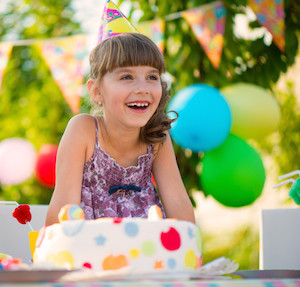 Little girl at a birthday party laughing and sitting in front of a cake, wearing a party hat and balloons in the background